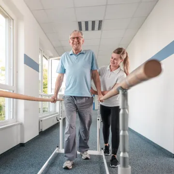 Ein Mann, der sich auf einem Gehbarren festhält und eine Physiotherapeutin, die ihn dabei unterstützt. 