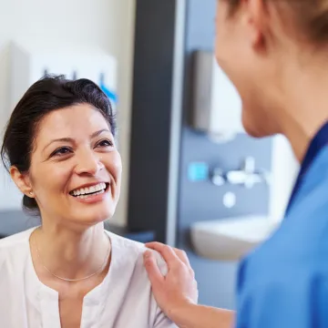 © Adobe Stock Eine Ärztin in blauem Gewand legt ihre Hand auf die Schulter einer dunkelhaarigen Patientin