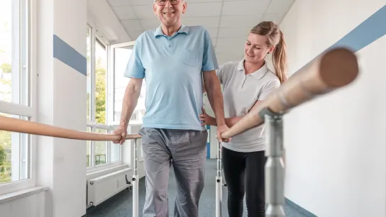 Ein Mann, der sich auf einem Gehbarren festhält und eine Physiotherapeutin, die ihn dabei unterstützt. 
